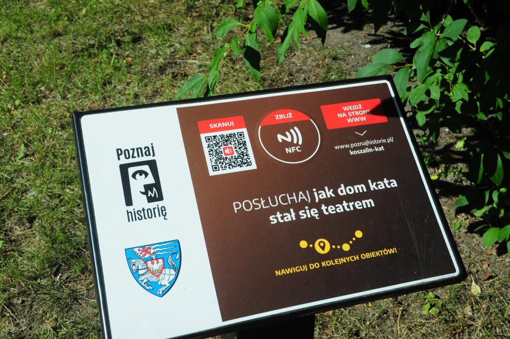 Tablica informacyjna z kodem QR, symbolem NFC i napisem „Posłuchaj, jak dom kata stał się teatrem”; poniżej znajduje się informacja o możliwości nawigacji do kolejnych obiektów (kliknięcie powiększy zdjęcie).