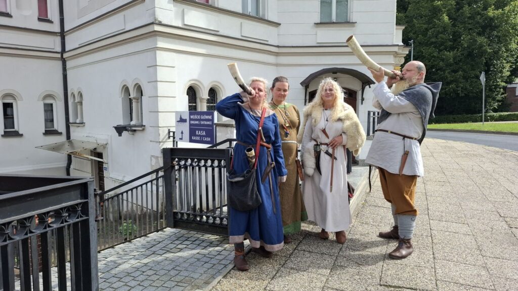 Cztery osoby w strojach inspirowanych średniowieczem stoją przed budynkiem muzeum; dwie osoby trzymają przy ustach rogi do grania; w tle widoczne jest wejście do muzeum i oznaczenie kasy (kliknięcie powiększy zdjęcie).