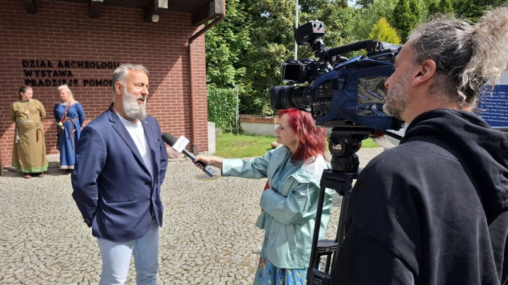Rozmowa przed budynkiem muzeum; mężczyzna udziela wywiadu dziennikarce, obok operator kamery nagrywa rozmowę; w tle widoczny budynek działu archeologii i wejście na wystawę stałą "Pradzieje Pomorza" (kliknięcie powiększy zdjęcie).