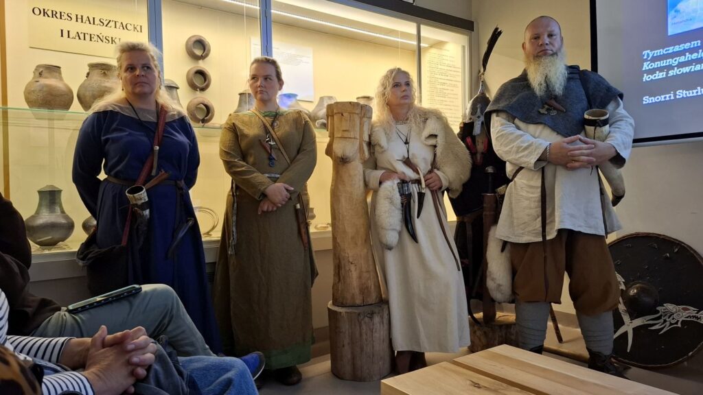 Grupa osób w strojach historycznych stoi przed uczestnikami wydarzenia; w tle widoczne są muzealne gabloty z zabytkami archeologicznymi oraz elementy wystawy poświęconej historii średniowiecza (kliknięcie powiększy zdjęcie).