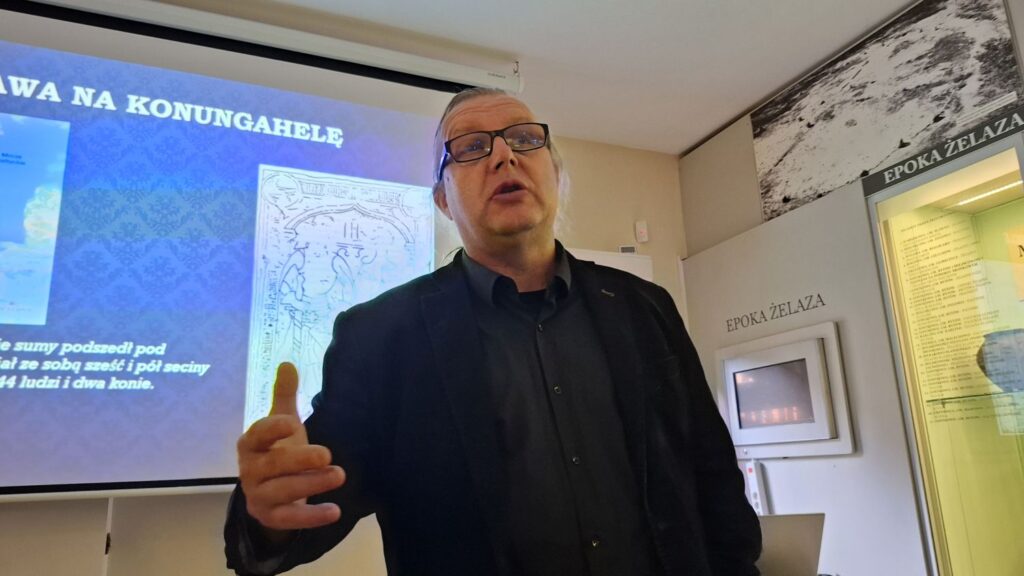 Mężczyzna prowadzący prelekcję stoi przed ekranem prezentacyjnym; na ekranie widoczny jest slajd dotyczący wyprawy na Konungahellę; w tle znajdują się muzealne gabloty i plansze wystawowe (kliknięcie powiększy zdjęcie).