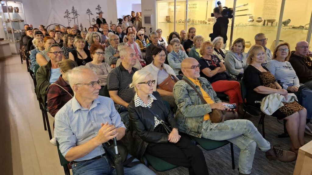 Liczna grupa uczestników wydarzenia siedzi w sali muzealnej; część osób patrzy w stronę prowadzącego i ekranu z prezentacją; w tle widoczne są gabloty oraz dekoracja ścienna z sylwetkami drzew (kliknięcie powiększy zdjęcie).
