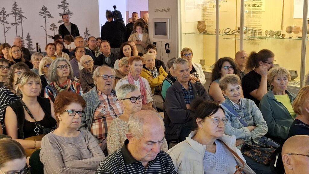 Uczestnicy wydarzenia siedzą w muzealnej sali podczas prelekcji; w tle widoczne są gabloty z zabytkami i plansze wystawowe; część osób skupia uwagę na prezentacji (kliknięcie powiększy zdjęcie).