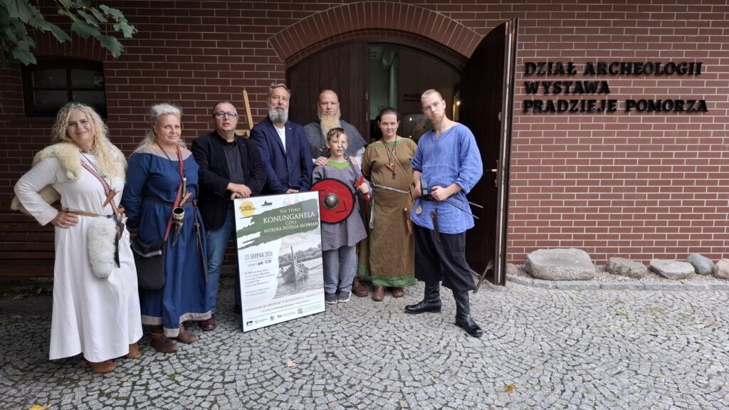 Grupa osób w strojach inspirowanych średniowieczem pozuje przed wejściem do budynku wystawy archeologicznej "Pradzieje Pomorza"; uczestnicy trzymają rekwizyty nawiązujące do epoki, a przed nimi znajduje się plakat wydarzenia dotyczącego wyprawy na Konungahellę (kliknięcie powiększy zdjęcie).