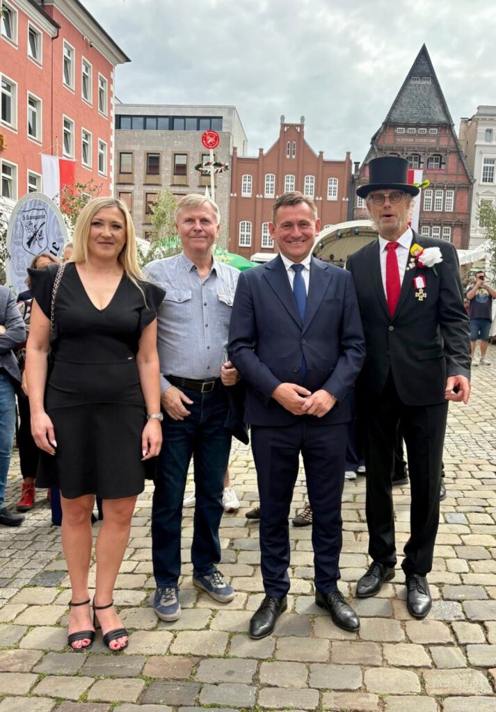 Cztery osoby pozują na brukowanym placu w centrum miasta; w tle widoczne są zabytkowe kamienice, wieża kościoła i uczestnicy miejskiego wydarzenia (kliknięcie powiększy zdjęcie).