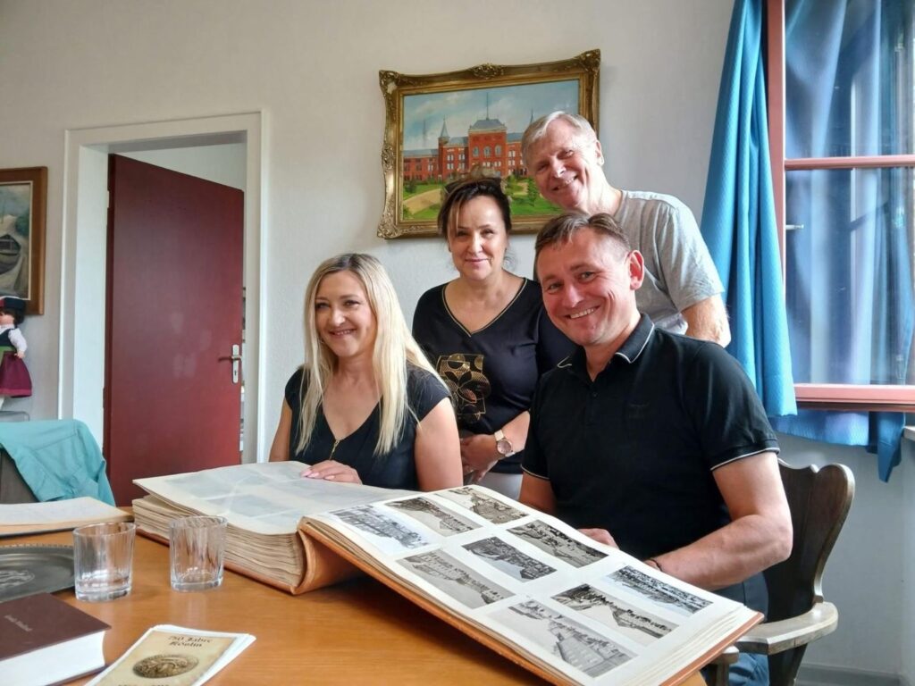 Cztery osoby pozują przy stole z otwartą księgą zawierającą archiwalne fotografie i dokumenty; w tle widoczne są obrazy przedstawiające zabudowę miejską (kliknięcie powiększy zdjęcie).