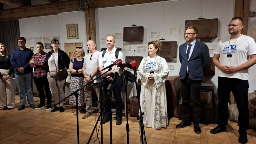Grupa osób podczas konferencji prasowej uczestnicy stoją przy mikrofonach i pozują do zdjęcia w sali wystawy czasowej; w tle widać eksponaty wystawy (kliknięcie powiększy zdjęcie).