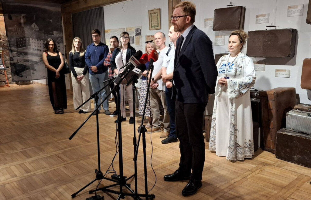 Mężczyzna stoi przed mikrofonem i przemawia podczas konferencji prasowej; za nim stoją pozostali uczestnicy; w tle widać eksponaty wystawy (kliknięcie powiększy zdjęcie).