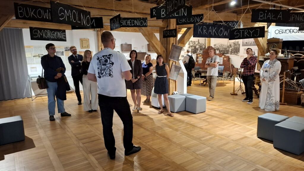 Uczestnicy wydarzenia zgromadzeni w sali muzealnej; na ścianach widoczne tabliczki z nazwami miejscowości, a na środku sali stoją osoby prowadzące spotkanie (kliknięcie powiększy zdjęcie).