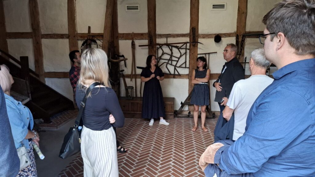 Grupa osób podczas zwiedzania muzeum; przewodniczka opowiada o ekspozycji w sali z drewnianą konstrukcją ścian i podłogą ułożoną we wzór cegieł; na ścianach wiszą różne metalowe narzędzia rolnicze (kliknięcie powiększy zdjęcie).
