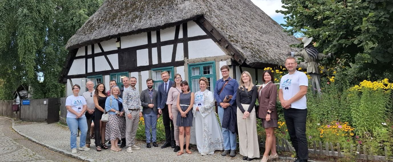 Grupa kilkunastu osób, uczestników konferencji prasowej i oprowadzania, pozuje do wspólnego zdjęcia przed zabytkowym budynkiem o konstrukcji szachulcowej, z dachem krytym strzechą; w tle drzewa o ogród (kliknięcie powiększy zdjęcie).