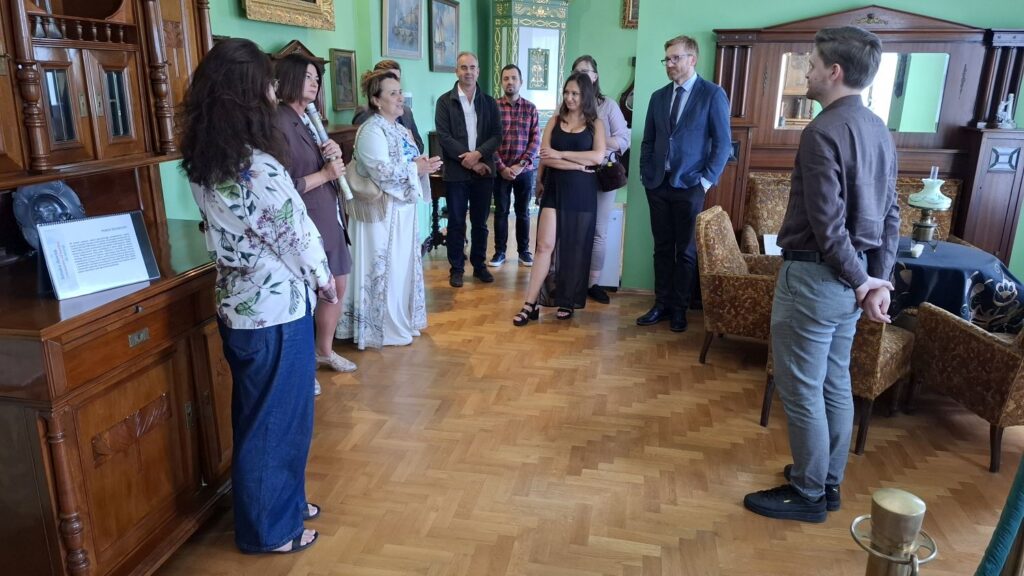 Grupa osób podczas zwiedzania muzealnej sali; przewodnik przedstawia ekspozycję, a w pomieszczeniu widoczne są zabytkowe meble i wyposażenie wnętrza (kliknięcie powiększy zdjęcie).
