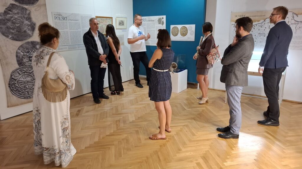 Grupa osób podczas zwiedzania wystawy historycznej; uczestnicy oglądają plansze z informacjami, mapami i reprodukcjami zabytków, a przewodnik opowiada o ekspozycji (kliknięcie powiększy zdjęcie).