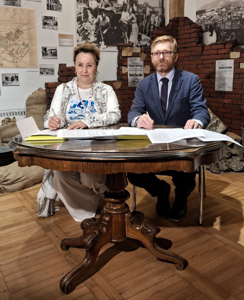 Podpisanie umowy przy zabytkowym drewnianym stole; kobieta i mężczyzna siedzą przy stole i składają podpisy na dokumentach, w tle widoczna wystawa muzealna z fotografiami, mapami i fragmentem ceglanego muru (kliknięcie powiększy zdjęcie).