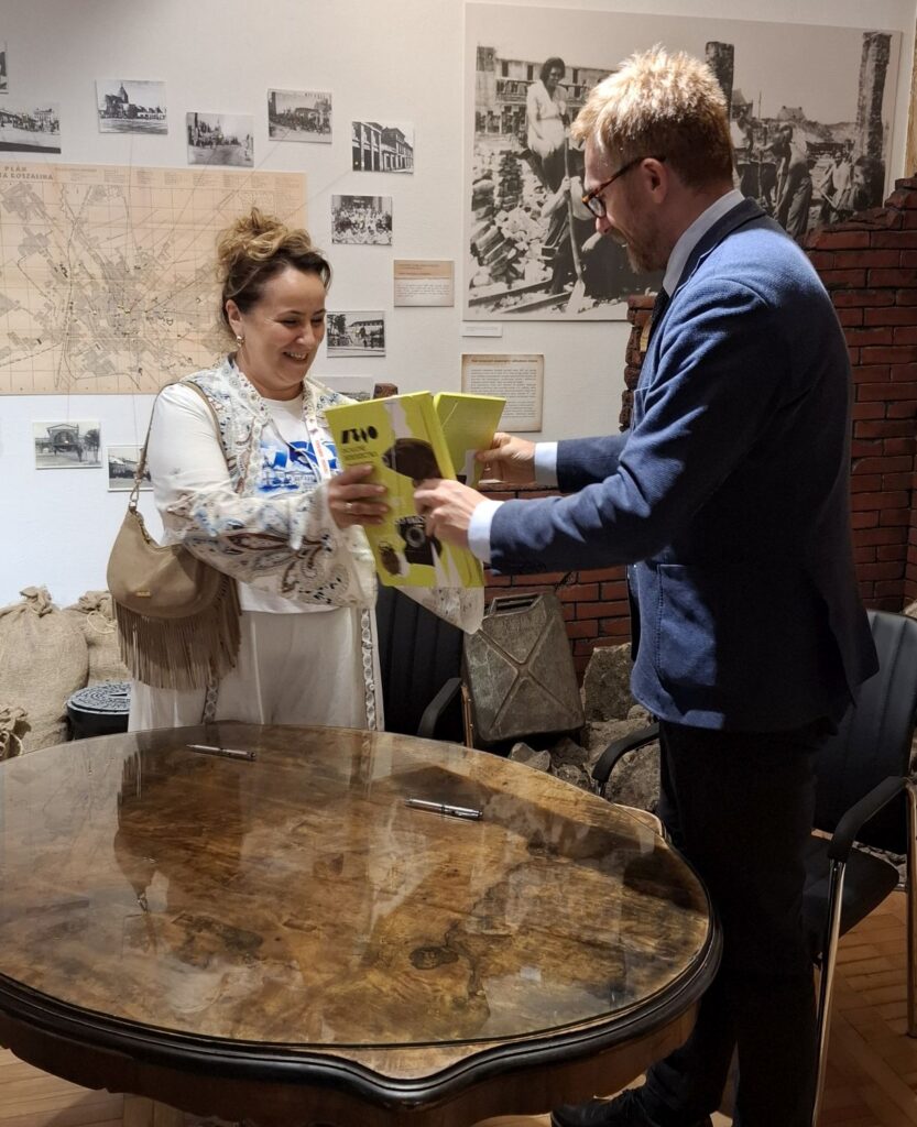 Kobieta i mężczyzna przekazują sobie żółtą teczkę, w tle widoczna wystawa muzealna z fotografiami, mapami i fragmentem ceglanego muru (kliknięcie powiększy zdjęcie).
