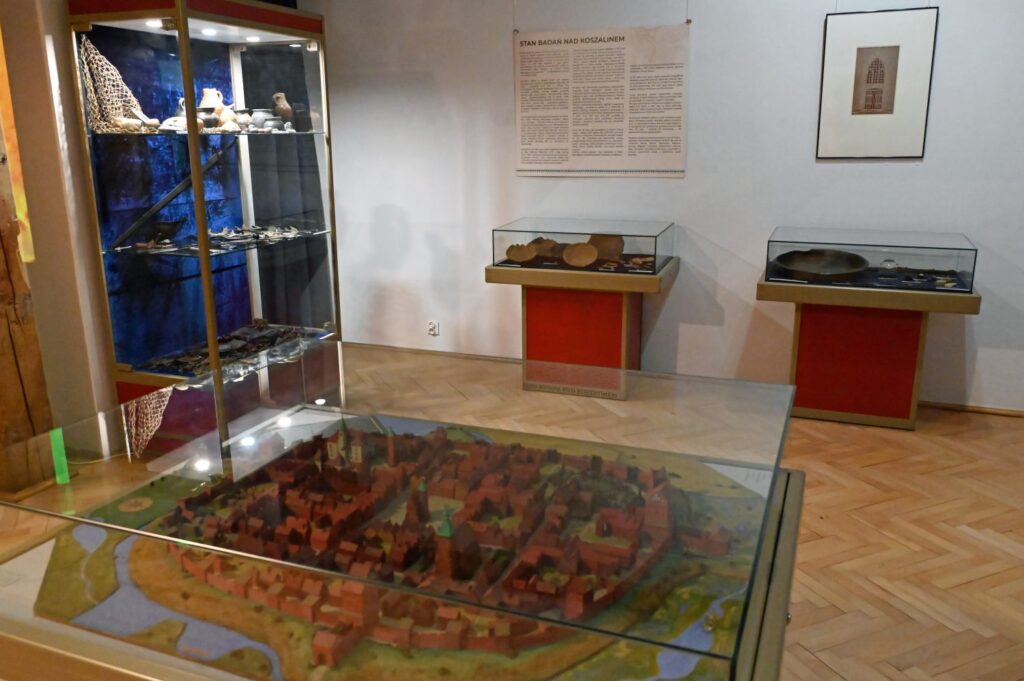 Makieta średniowiecznego Koszalina prezentowana w gablocie na tle ekspozycji z zabytkami archeologicznymi i planszami historycznymi (kliknięcie powiększy zdjęcie).