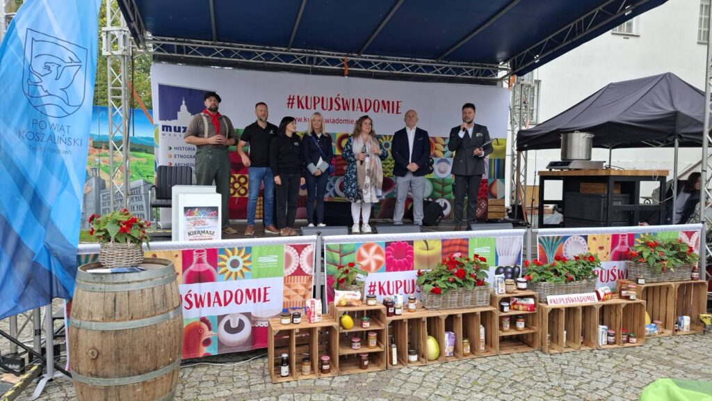 Scena plenerowa podczas festynu; na scenie stoi grupa osób, a przed nią znajduje się ekspozycja produktów spożywczych ustawionych na drewnianych skrzynkach, beczka i dekoracje z kwiatami (kliknięcie powiększy zdjęcie).