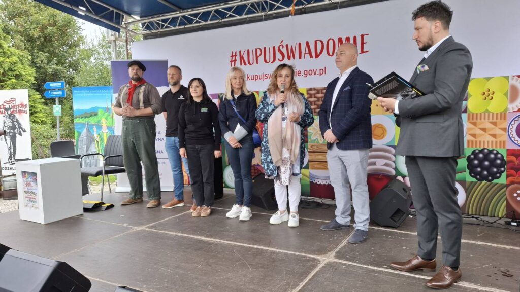 Uczestnicy wydarzenia stoją na scenie przed kolorowym banerem z napisem "#KUPUJŚWIADOMIE”; kobieta przemawia do zgromadzonych osób, a mężczyzna po prawej trzyma w rękach publikację (kliknięcie powiększy zdjęcie).