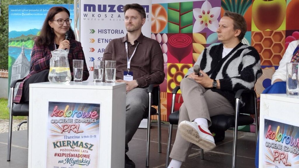 Paneliści podczas rozmowy na scenie; trzy osoby siedzą na krzesłach, a przed nimi ustawione są stoliki, na których ustawione są szklanki i woda do picia (kliknięcie powiększy zdjęcie).