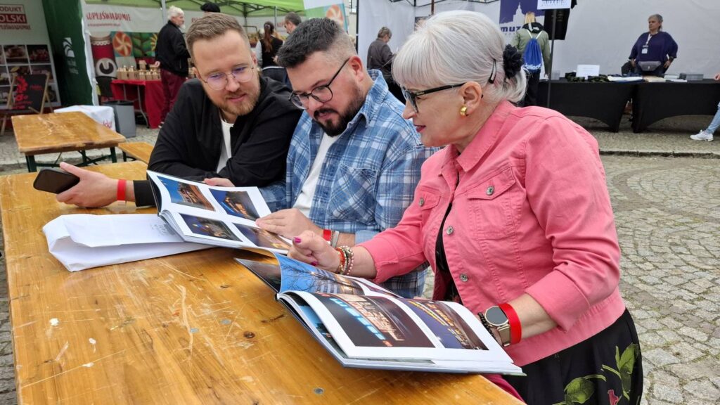 Trzy osoby przeglądają wspólnie album przy drewnianym stole; oglądają fotografie i rozmawiają podczas wydarzenia plenerowego (kliknięcie powiększy zdjęcie).