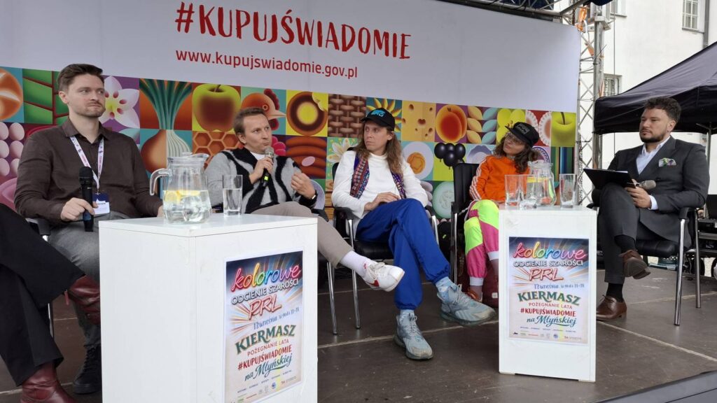 Uczestnicy panelu dyskusyjnego na scenie; cztery osoby siedzą przy stolikach z publikacjami i materiałami promocyjnymi (kliknięcie powiększy zdjęcie).
