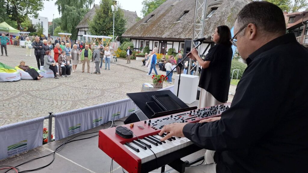 Muzyk gra na czerwonym keyboardzie na scenie, towarzysząc osobie prowadzącej wydarzenie; w tle widać zgromadzoną publiczność oraz zabudowania skansenu (kliknięcie powiększy zdjęcie).