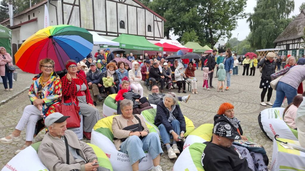 Publiczność siedzi na kolorowych pufach przed sceną; wśród widzów wyróżniają się osoby z wielobarwnym parasolem i kolorowymi strojami, a w tle znajdują się namioty i zabytkowe zabudowania (kliknięcie powiększy zdjęcie).