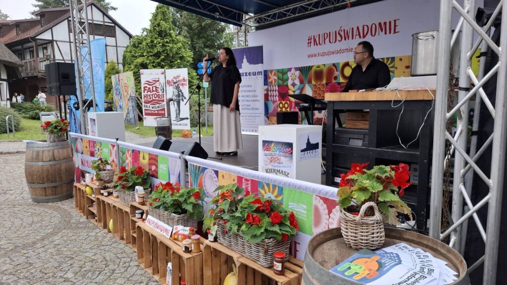 Scena plenerowa podczas wydarzenia; na scenie stoi kobieta z mikrofonem, a przed nią znajduje się dekoracja z kwiatami, drewnianymi skrzynkami i materiałami promocyjnymi (kliknięcie powiększy zdjęcie).
