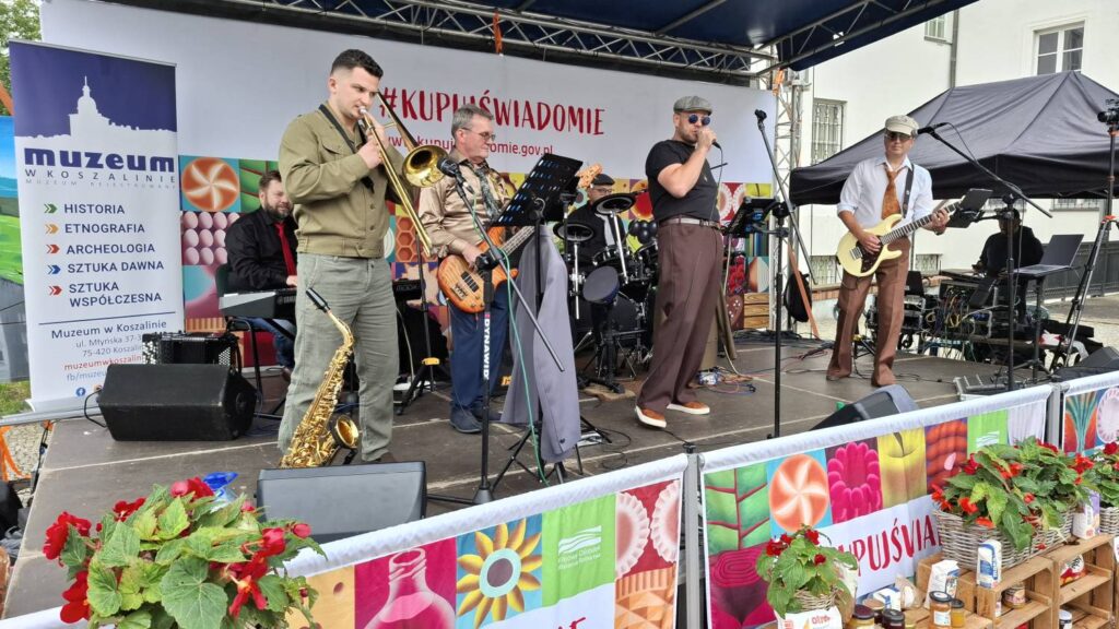 Zespół muzyczny występuje na plenerowej scenie; muzycy grają na saksofonie, puzonie, gitarach, perkusji i innych instrumentach, a scenę zdobią kolorowe grafiki i kwiaty (kliknięcie powiększy zdjęcie).