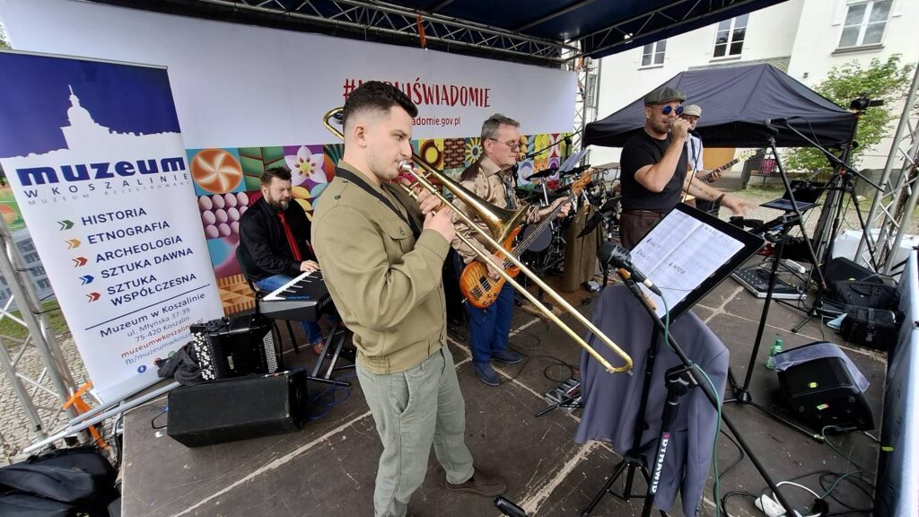 Muzycy podczas koncertu na plenerowej scenie; na pierwszym planie instrumentalista gra na puzonie, a w tle widoczni są pozostali członkowie zespołu z gitarami i mikrofonami (kliknięcie powiększy zdjęcie).