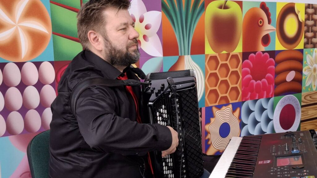 Muzyk gra na akordeonie i keyboardzie; za nim znajduje się kolorowa, geometryczna dekoracja sceny z motywami owoców, kwiatów i abstrakcyjnych wzorów (kliknięcie powiększy zdjęcie).