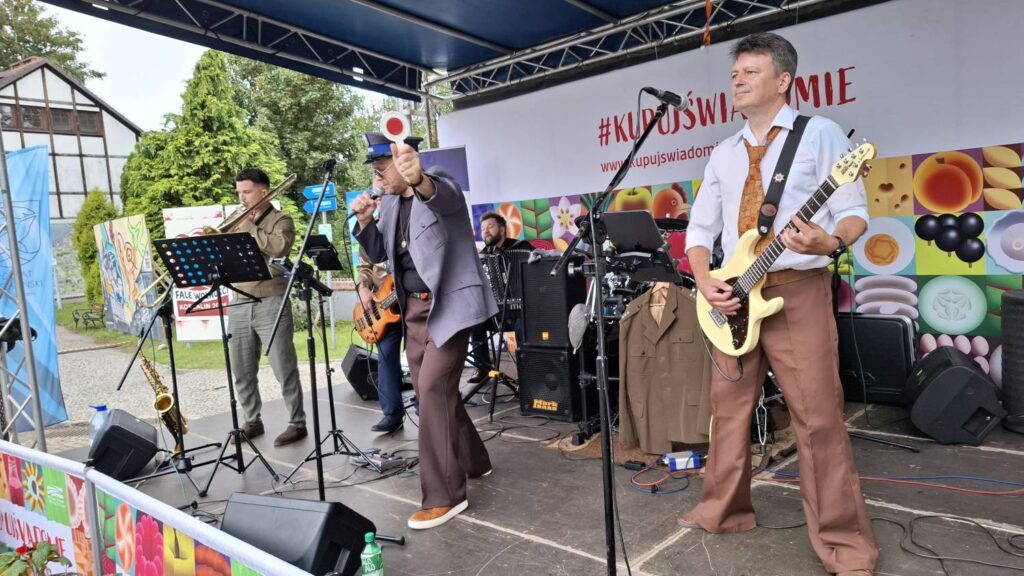 Zespół występuje na scenie; na pierwszym planie gitarzysta gra na jasnej gitarze elektrycznej, obok niego wokalista trzyma mikrofon, a w tle widoczni są pozostali muzycy i instrumenty (kliknięcie powiększy zdjęcie).