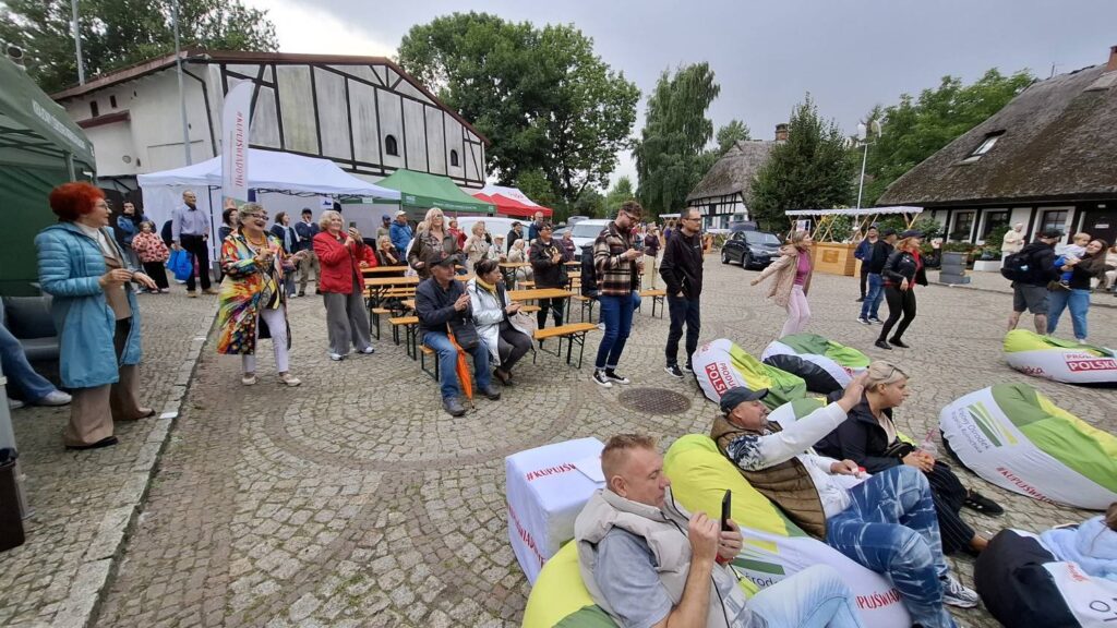 Publiczność słucha koncertu na brukowanym dziedzińcu; część osób siedzi na kolorowych pufach, inni stoją lub tańczą, a w tle znajdują się scena, namioty i zabytkowe zabudowania (kliknięcie powiększy zdjęcie).
