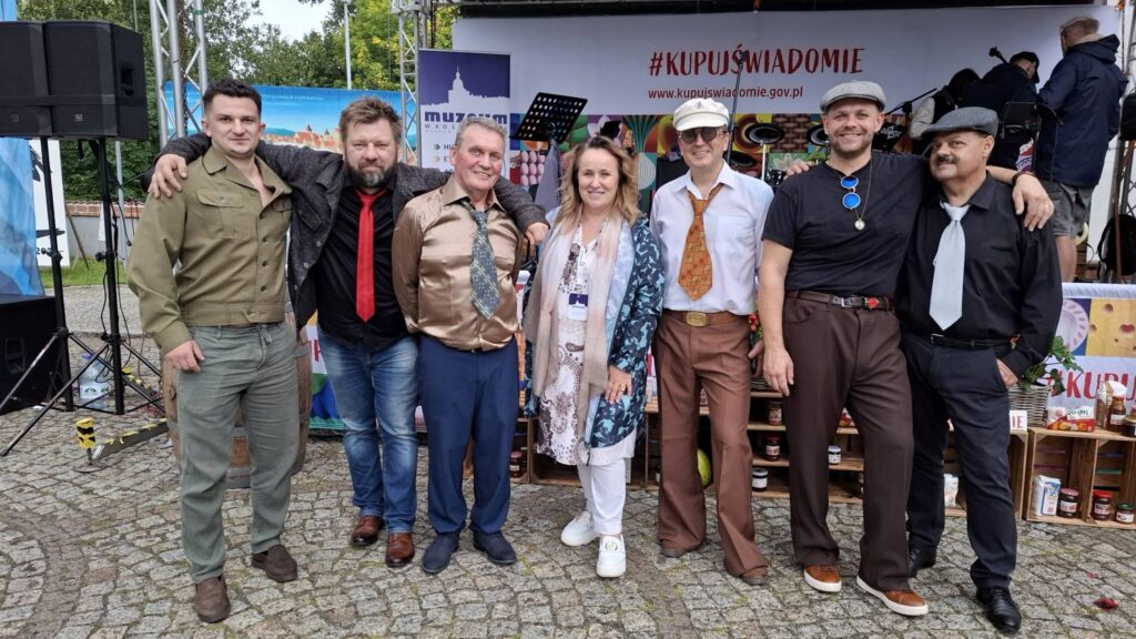 Członkowie zespołu muzycznego pozują wspólnie na scenie po występie; grupa stoi przed kolorową dekoracją, wśród muzyków widoczne są gitary, mikrofony i inne elementy wyposażenia sceny (kliknięcie powiększy zdjęcie).