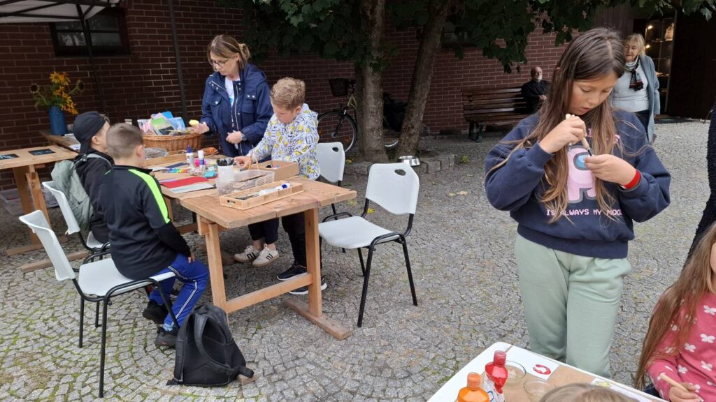 Dzieci uczestniczą w zajęciach plastycznych na świeżym powietrzu; przy drewnianych stołach rysują i wykonują prace plastyczne, korzystając z przygotowanych materiałów (kliknięcie powiększy zdjęcie).