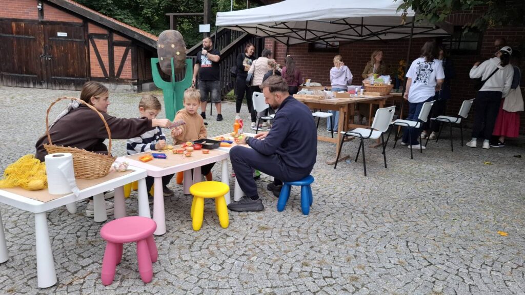 Dzieci i dorośli biorą udział w warsztatach plastycznych na muzealnym dziedzińcu; uczestnicy pracują przy stołach, a obok znajdują się kolorowe dziecięce stołeczki (kliknięcie powiększy zdjęcie).