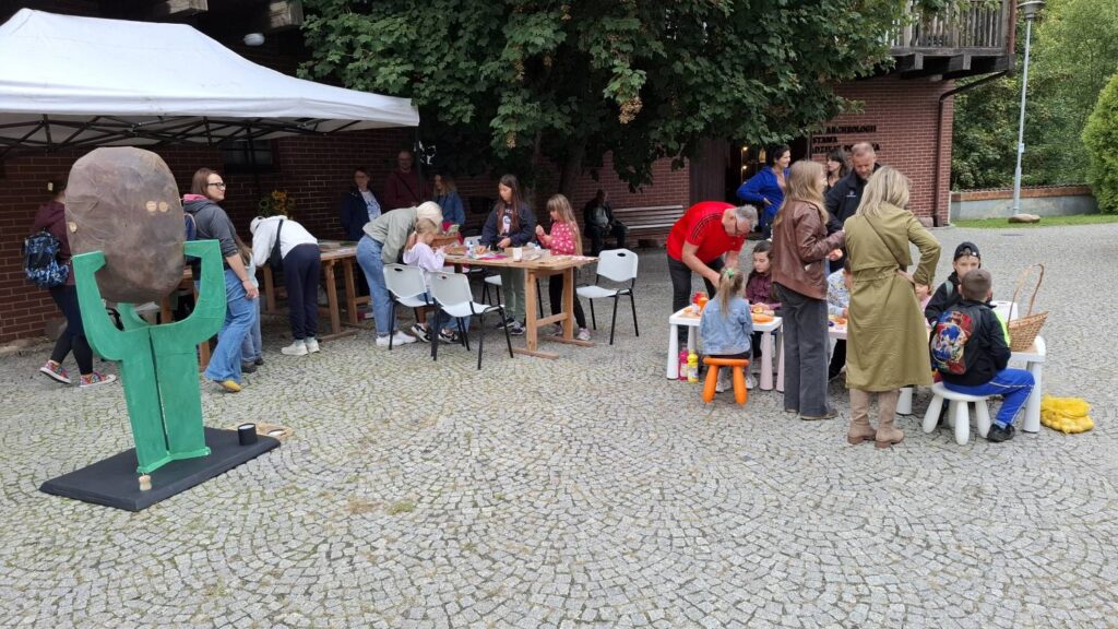 Uczestnicy wydarzenia spędzają czas na muzealnym dziedzińcu; część osób bierze udział w zajęciach przy stołach, a na pierwszym planie znajduje się duża zielona rzeźba z kamienną głową (kliknięcie powiększy zdjęcie).