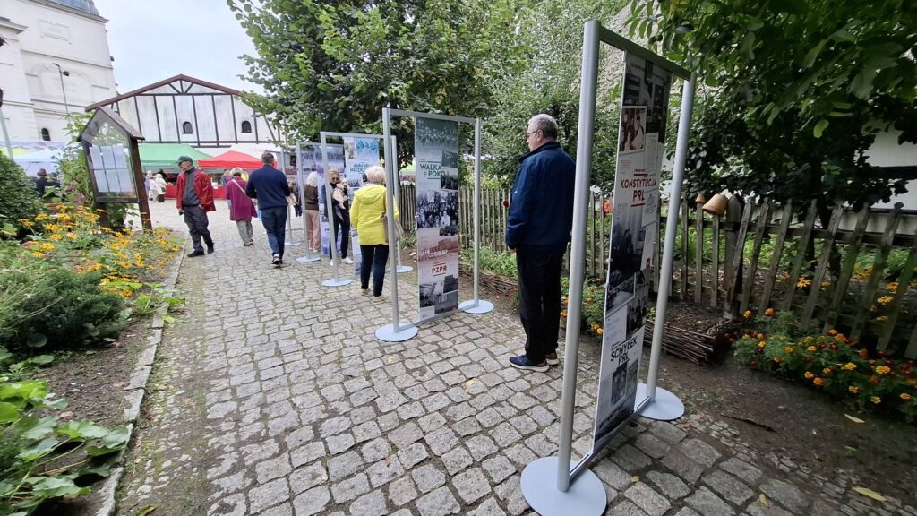 Zwiedzający oglądają plenerową wystawę historyczną; plansze z archiwalnymi fotografiami i materiałami dotyczącymi PRL ustawiono wzdłuż brukowanej ścieżki wśród zieleni (kliknięcie powiększy zdjęcie).