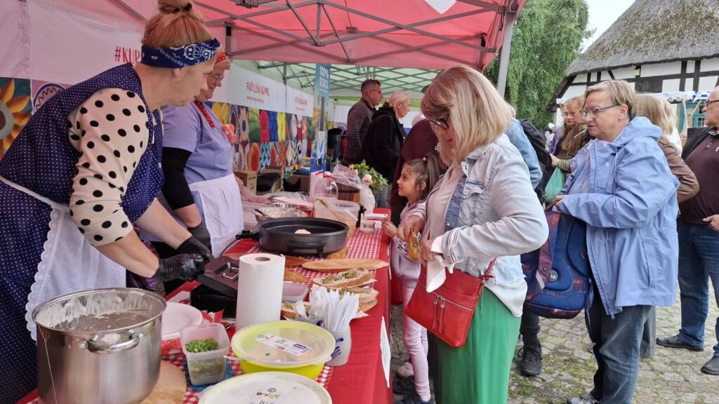 Kobiety przygotowują i serwują potrawy przy stoisku gastronomicznym; na stole znajdują się garnki, miski, talerze oraz przygotowane jedzenie (kliknięcie powiększy zdjęcie).
