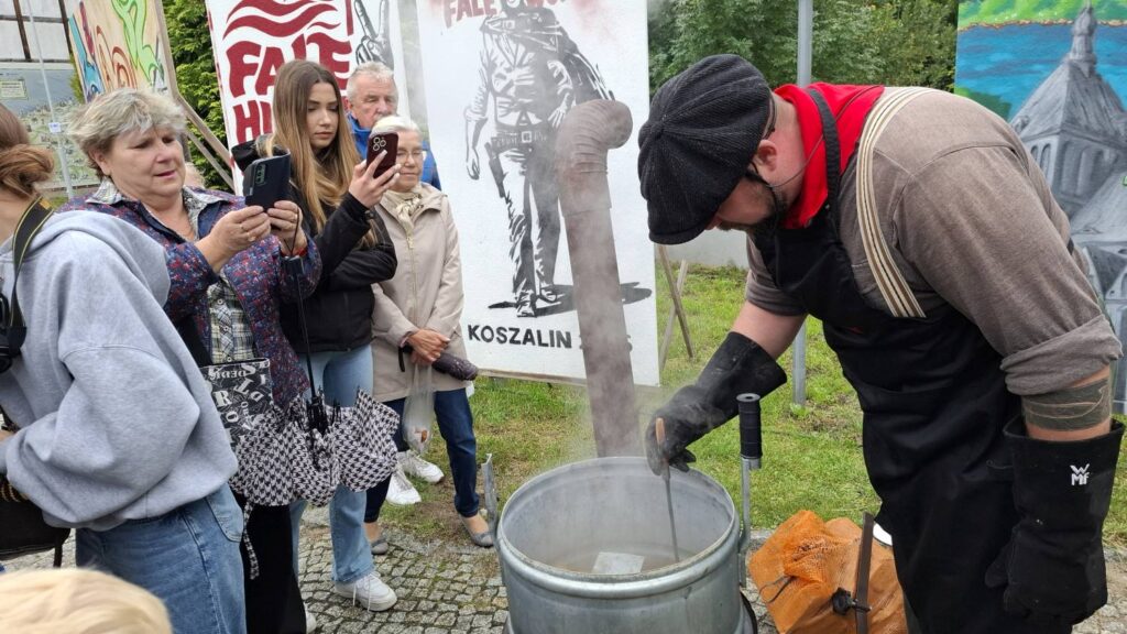 Mężczyzna w czapce i fartuchu przygotowuje potrawę w dużym metalowym kotle; wokół zgromadzili się uczestnicy obserwujący gotowanie, a nad kotłem unosi się para (kliknięcie powiększy zdjęcie).
