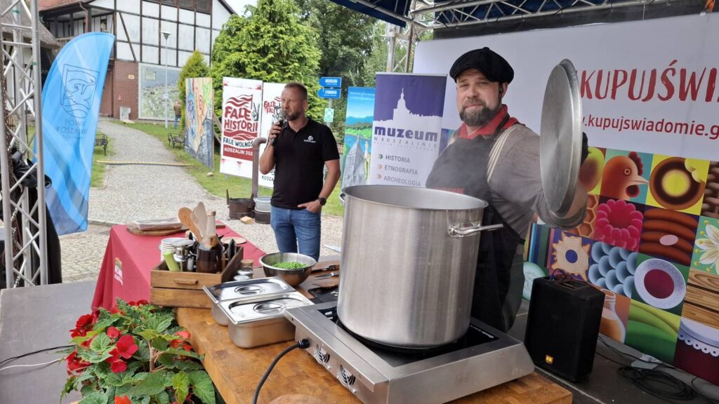 Mężczyzna prezentuje duży metalowy garnek podczas pokazu kulinarnego; obok znajdują się przygotowane składniki i przybory kuchenne, a w tle widoczna jest scena wydarzenia (kliknięcie powiększy zdjęcie).