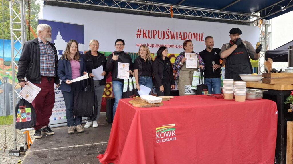 Grupa uczestników wydarzenia pozuje na scenie z dyplomami i materiałami promocyjnymi; przed nimi znajduje się stół z czerwonym obrusem i naczyniami używanymi podczas pokazu kulinarnego (kliknięcie powiększy zdjęcie).