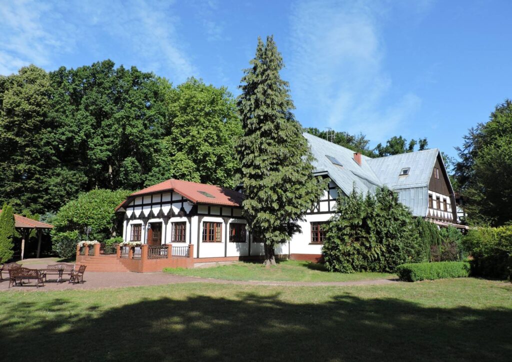 Zabytkowy budynek o konstrukcji szachulcowej, otoczony zielenią; przed budynkiem znajduje się taras z drewnianymi meblami ogrodowymi (kliknięcie powiększy zdjęcie).