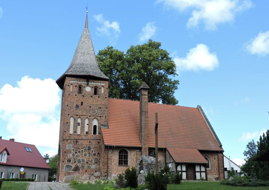 Zabytkowy kościół, zbudowany z czerwonej cegły i kamieni, z charakterystyczną wysoką wieżą i stromym dachem, otoczony zielenią i zabudową mieszkalną (kliknięcie powiększy zdjęcie).