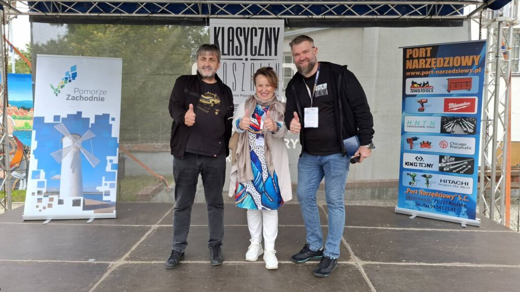 Dwóch mężczyzn i jedna kobieta, przedstawiciele organizatorów wydarzenia motoryzacyjnego, pozują na scenie do wspólnego zdjęcia; za nimi widoczne są banery promocyjne i scena plenerowa (kliknięcie powiększy zdjęcie).