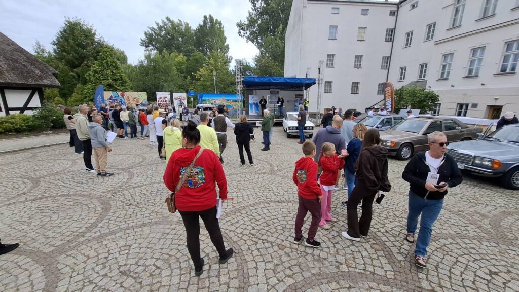 Grupa uczestników spaceruje na dziedzińcu podczas zlotu samochodów zabytkowych; wokół znajdują się klasyczne auta oraz zabudowania muzeum (kliknięcie powiększy zdjęcie).