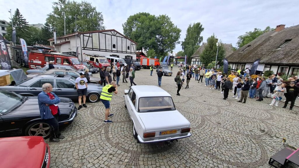 Uczestnicy wydarzenia oglądają zabytkowe samochody stojące na dziedzińcu muzeum; wzdłuż placu stoją zaparkowane pojazdy i zgromadzeni widzowie; w tle widoczne są zabudowania muzeum (kliknięcie powiększy zdjęcie).