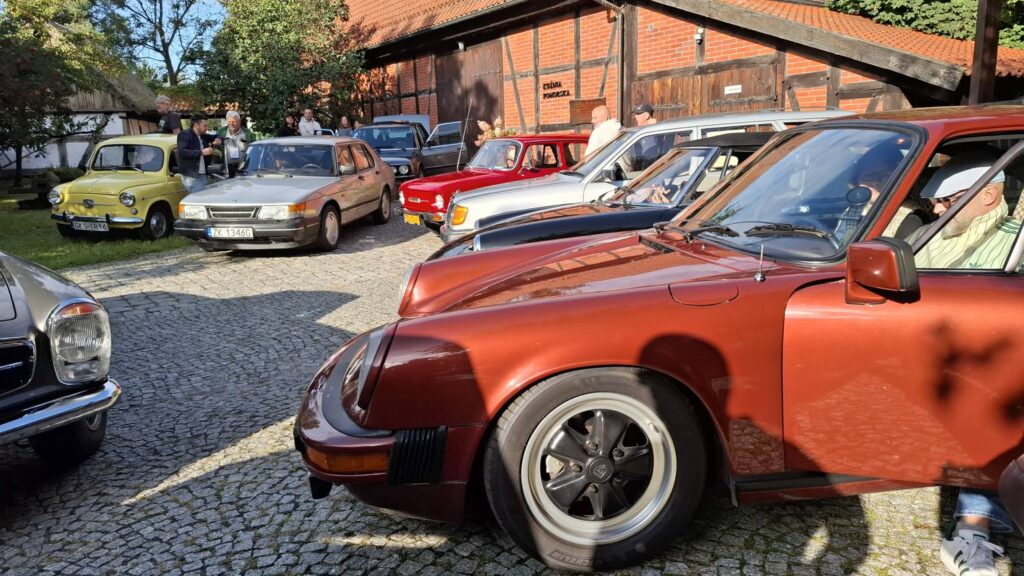 Rząd klasycznych samochodów zaparkowanych na brukowanym dziedzińcu; na pierwszym planie wyróżnia się pomarańczowo-brązowe auto sportowe, a dalej stoją m.in. żółte, czerwone i srebrne samochody (kliknięcie powiększy zdjęcie).