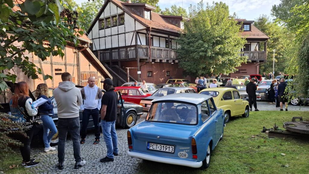 Niebieski klasyczny samochód znajduje się wśród innych zabytkowych aut zaparkowanych na terenie muzeum; w tle widoczni są uczestnicy oraz drewniana zabudowa (kliknięcie powiększy zdjęcie).