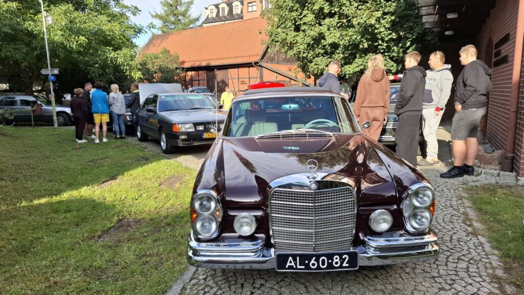 Zabytkowy samochód Mercedes w kolorze ciemnobrązowym stoi przed drewnianą zabudową; za nim zaparkowane są kolejne klasyczne auta, a wokół spacerują uczestnicy wydarzenia (kliknięcie powiększy zdjęcie).
